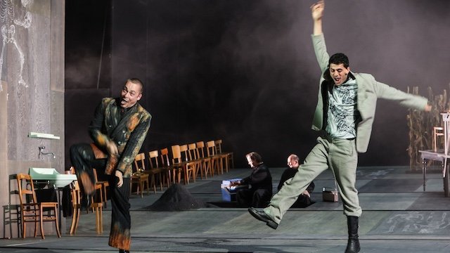 Zwei Männer stehen vorne auf einer Theaterbühne, machen Grimassen und ungewöhnliche Bewegungen. Im Hintergrund sitzen zwei Personen und links von Ihnen befindet sich eine Reihe von alten Holzstühlen.