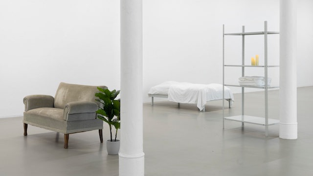 Ein Sessel, ein Bett und ein weißes Regal stehen in einem weiten, weißen Ausstellungsraum.