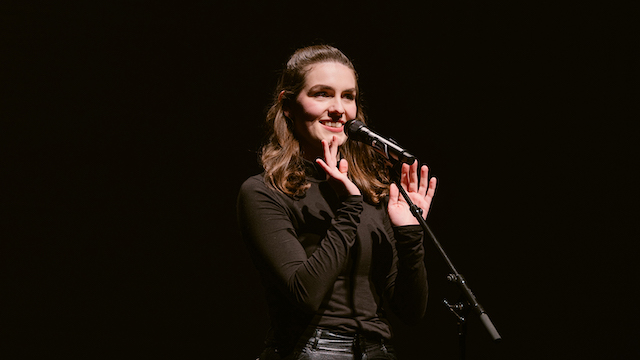 Eine junge Frau mit braunen Haaren steht in einem schwarzen Oberteil auf einer Bühne